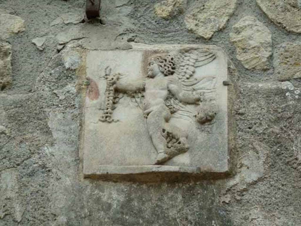 III.3 Herculaneum. May 2010. Detail of marble relief of cupid on wall above the pool.
The cupid is carrying in his hand, according to Jashemski, a purse filled with money. 
See Jashemski, W. F., 1993. The Gardens of Pompeii, Volume II: Appendices. New York: Caratzas. (p.259)
