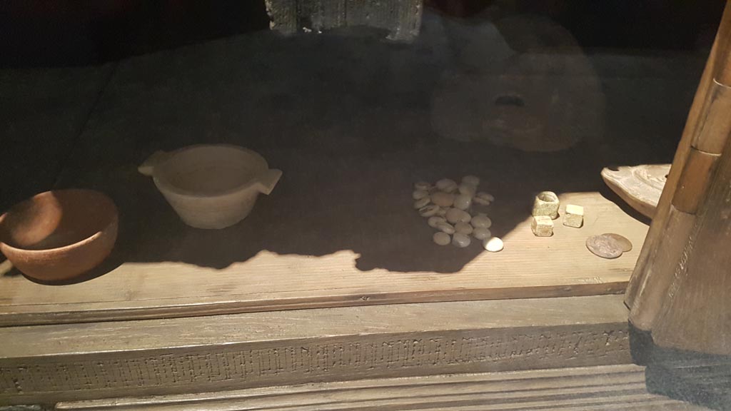 V.31 Herculaneum, August 2023. 
Wooden cupboard with items on display including loaded dice, on right. Photo courtesy of Maribel Velasco.

