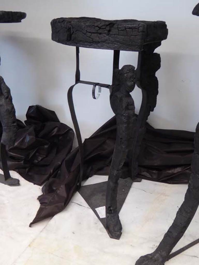 V.8 Herculaneum. September 2016. 
Carbonised wooden table with circular top and three legs decorated with human heads, or perhaps heads of cupids.
Photo courtesy of Michael Binns.
See De Carolis, E. (2007). Il mobile a Pompei ed Ercolano, letti tavoli sedie e armadi. Rome, “L’ERMA” de Bretsneider, (p.104) 
