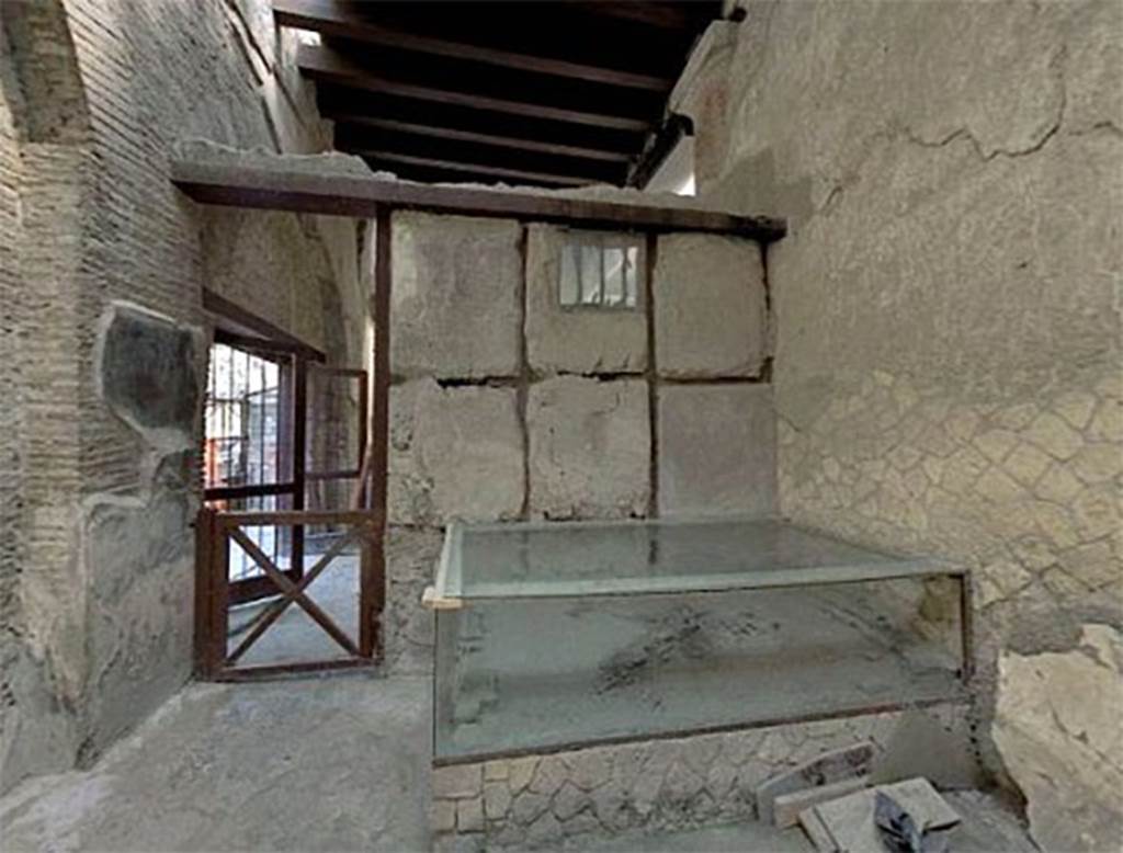 VI.21 Herculaneum. May 2006. Looking north across “caretaker’s room”, with remains of skeleton on its bed under a glass case.
Deiss wrote that this room had a door and a small rectangular window. 
The window is placed high and is barred. In the room was a wooden table of good quality and an elegant wooden bed. On the bed is the skeleton of a man who had thrown himself face downwards in defence against the volcanic gases…….   The supposition has been advanced that the little room was occupied by a caretaker or priest, and perhaps due to illness, he was unable to escape.
If the man was a caretaker, why should he have a temporary room adjoining the shrine? Why furniture of such high quality?  If a priest, why not a room of more impressive construction?  Why would the chief server at an emperor’s shrine be left behind on the flight of his fellows?  And if these living quarters, in spite of their hurried construction, were intended to be permanent, why no stove, no brazier for winter, no latrine?  Neither hypothesis takes account of the barred window or the evident despair in the man’s position.  It seems apparent that he could not escape, and no one unlocked the door…….  Whatever the reason for the man in the barred room of the Augustales, in the end he too paid with his life.  Perhaps someday a chance reference in literature will provide the key to that strange little room and its hopeless occupant.
See Deiss, J.J. 1968. Herculaneum: a city returns to the sun. UK, The History Book Club, (p.155).
